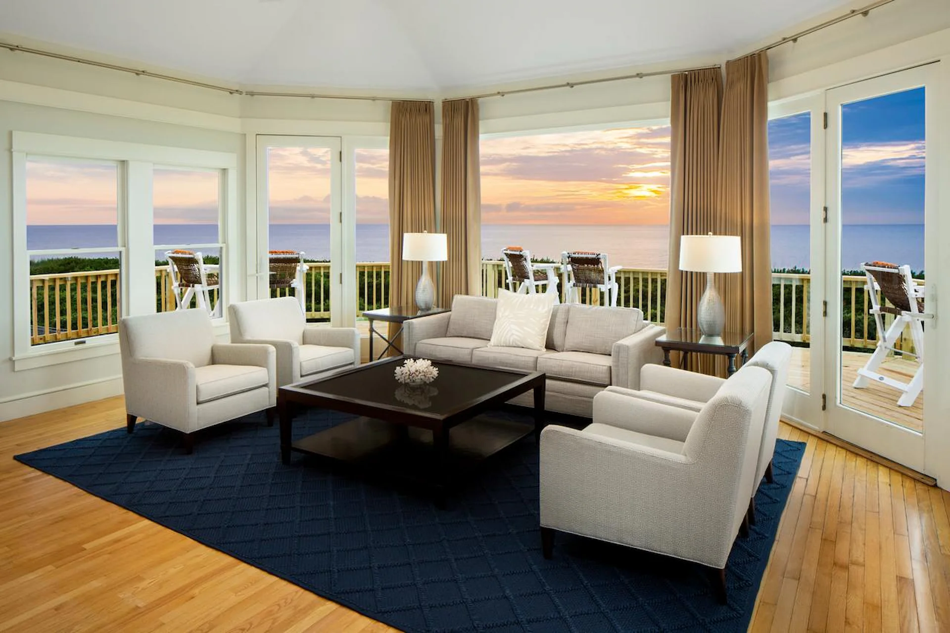 Elegant living room with ocean views, featuring neutral-toned furniture, a dark blue rug, and a sunset-lit terrace.