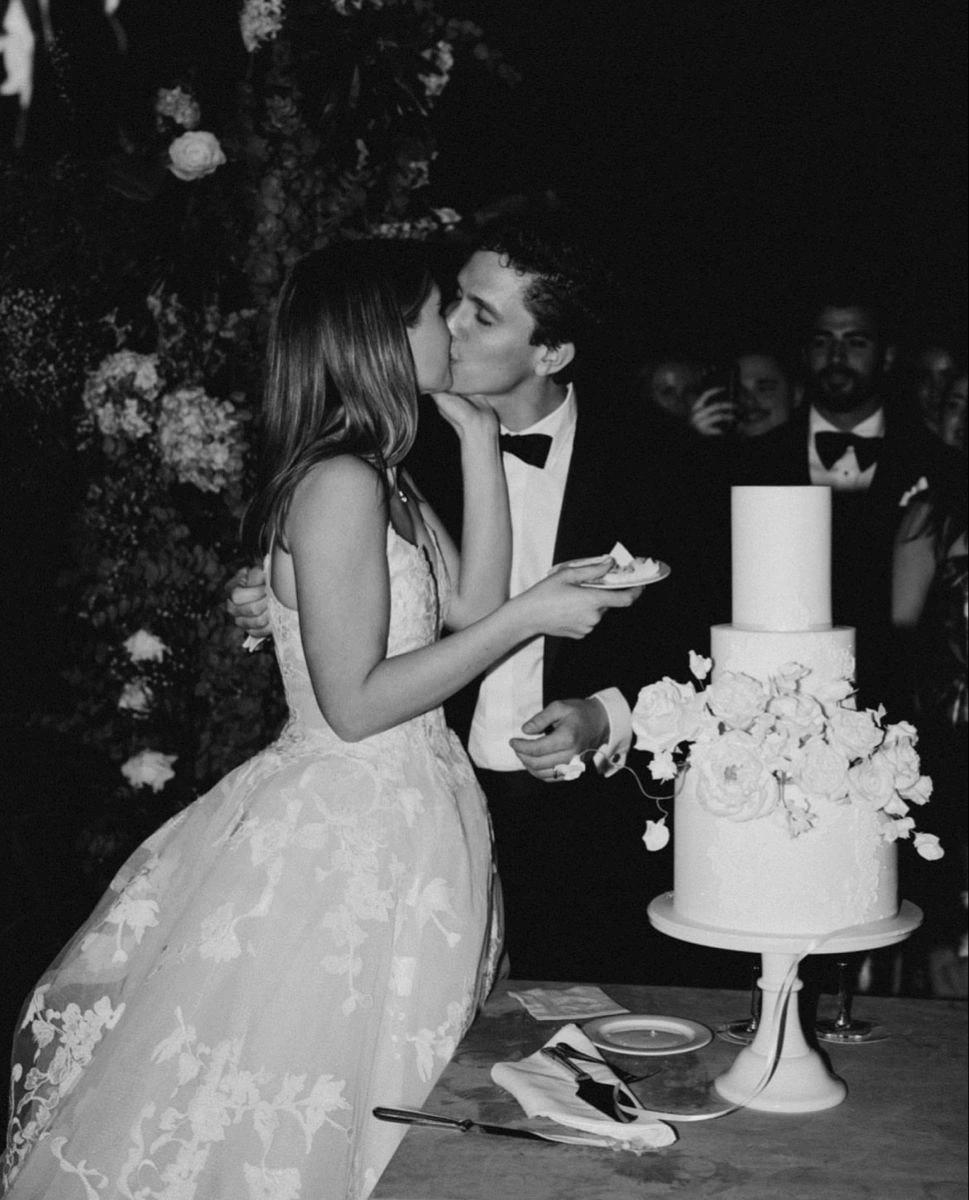 Black and white photo of a joyful newlywed couple kissing beside their wedding cake, with guests celebrating at Sanderling Hotel.